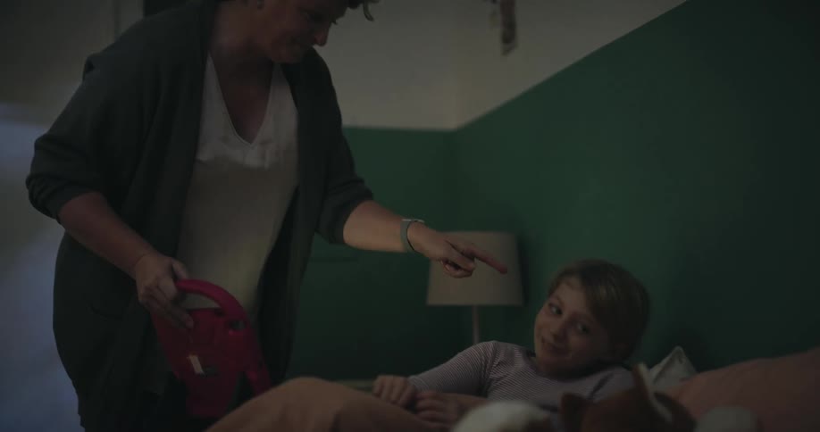 In a dimly lit bedroom a woman enters to take a tablet from a young girl sitting in bed then tucks her under the covers for sleep as they share a brief smiling conversation before the woman leaves the room