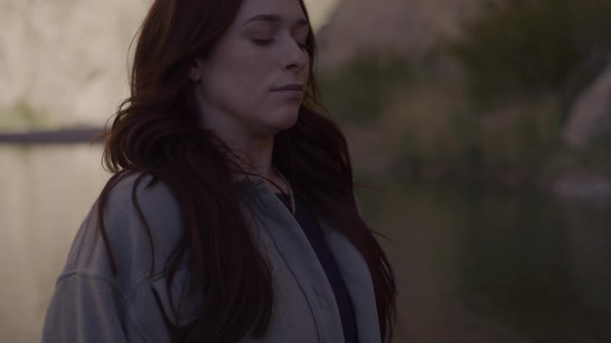 A young woman standing outdoors at midday scanning her surroundings before closing her eyes and taking a deep breath in a brief moment of meditation