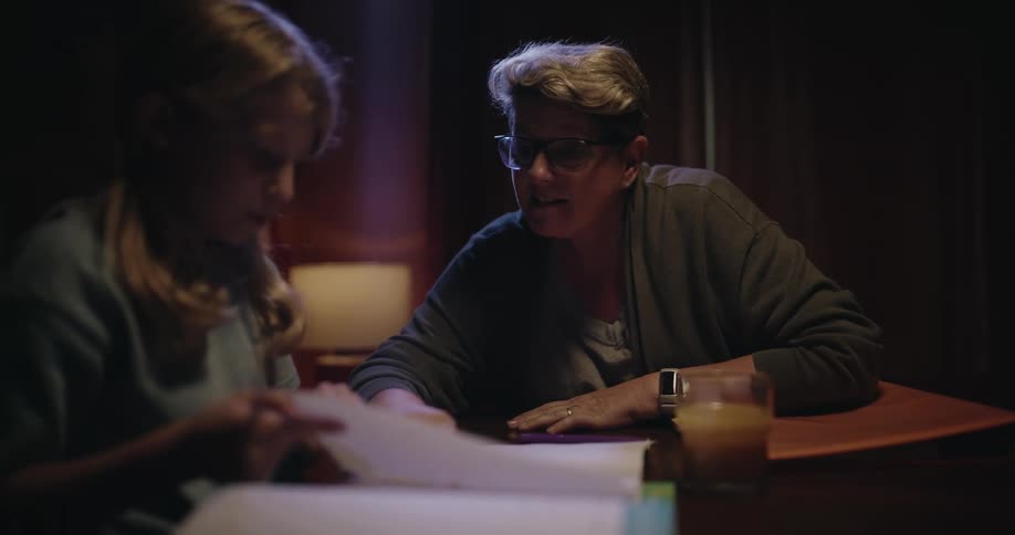 A young girl writes in a notebook with a pencil at a wooden table while a woman wearing glasses sits nearby and talks to her in a room with dim lighting and a glass of juice