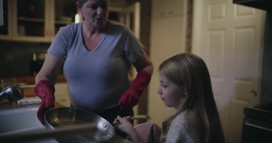 A woman with short grey hair wearing pink rubber gloves rinses a pan in a sink and hands it to a young girl who dries it with a cloth in a kitchen