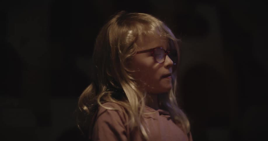 A child wearing glasses looking at a drawing in a dark room then letting it slip from their hands as it falls to the floor
