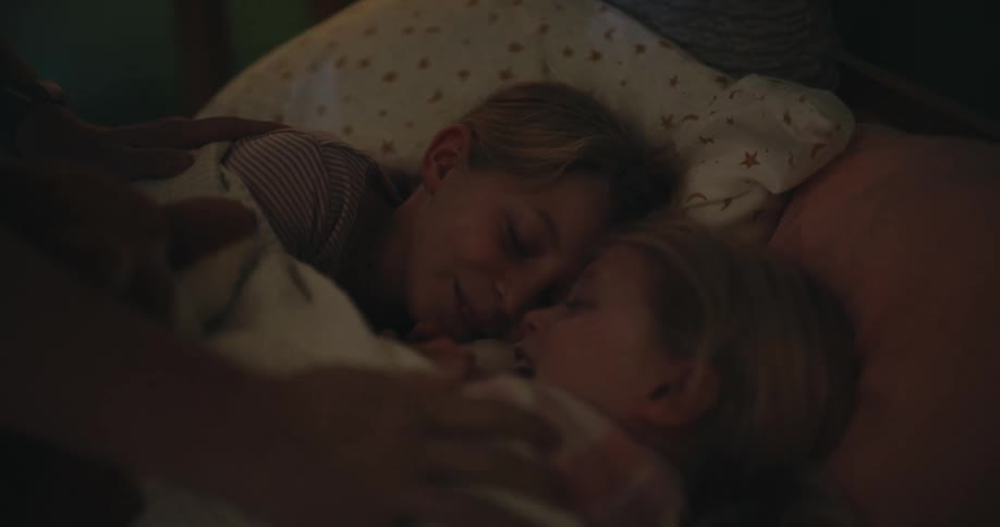 A close view of two young sisters sleeping peacefully together in a bed at night while a parent gently removes a brown teddy bear and tucks them in under a white blanket in a dimly lit bedroom