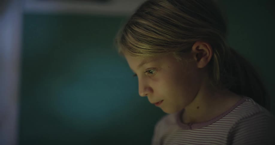 A young girl with long blonde hair inside a room looking down at a screen in front of her