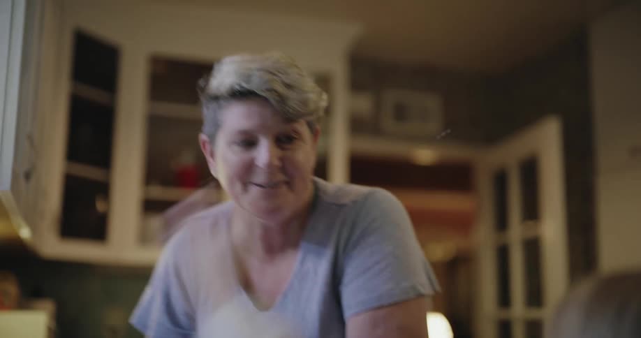 Adult woman wearing rubber gloves and washing dishes on the sink of a kitchen while a young girl playfully throws water on her face indoors during day time