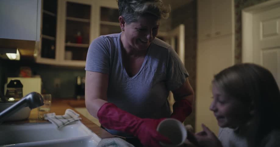 Person wearing red gloves washes dishes at the sink while smiling and engaging with a child