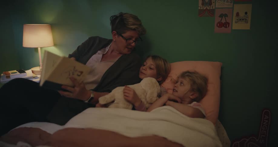 A mother sitting at the side of the bed reading bedtime stories to her two sleeping daughters then standing up afterward and gently taking off her glasses