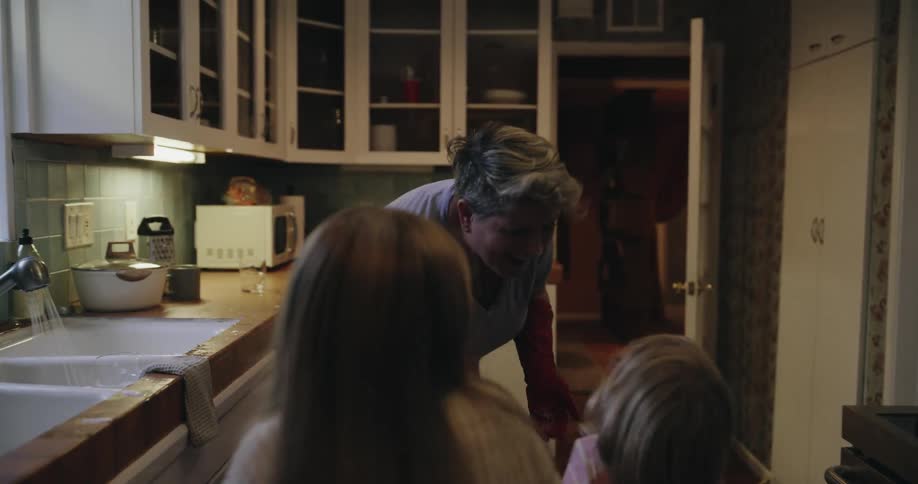 A mother in the kitchen doing the dishes while her two daughters stand beside her all three enjoying a fun lighthearted moment together