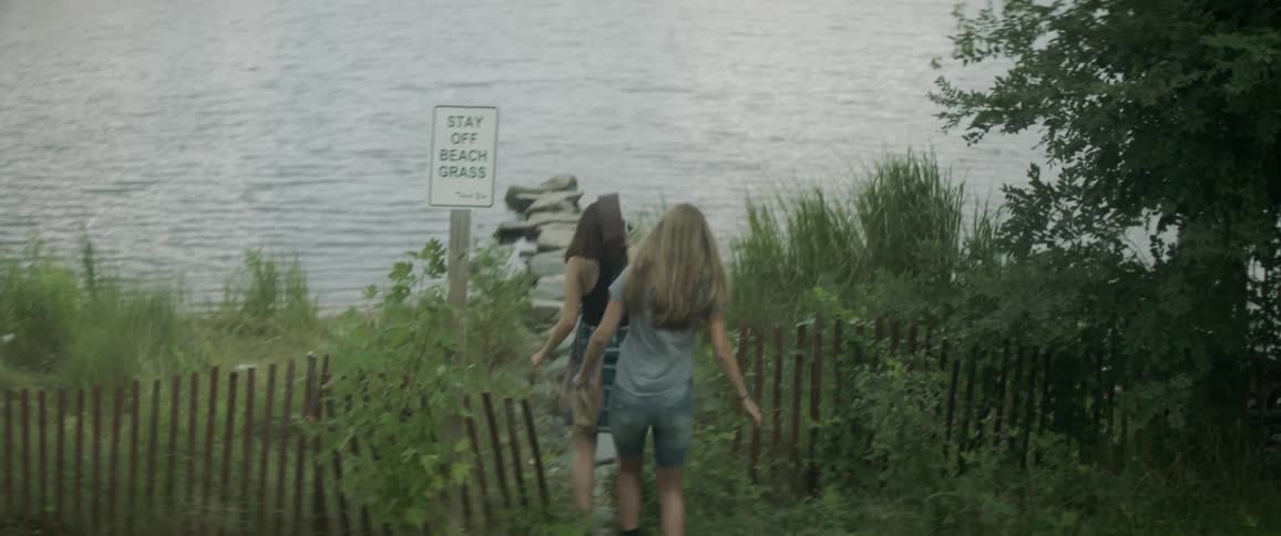 Two women walk towards the water on a beach, one of them carrying a guitar