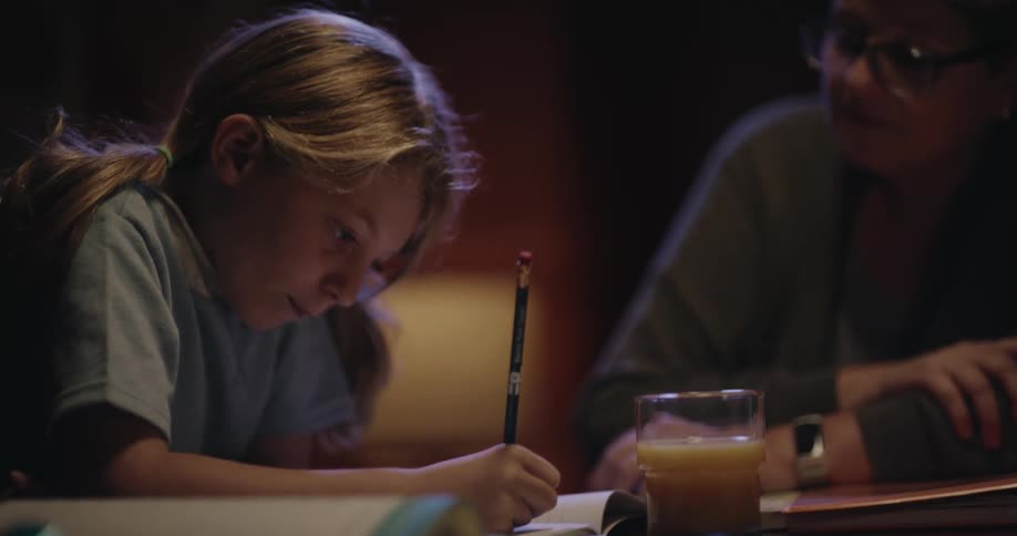A young girl with blonde hair tied in a ponytail focuses on writing in a notebook with a pencil while sitting at a table next to a woman wearing glasses in a dimly lit room