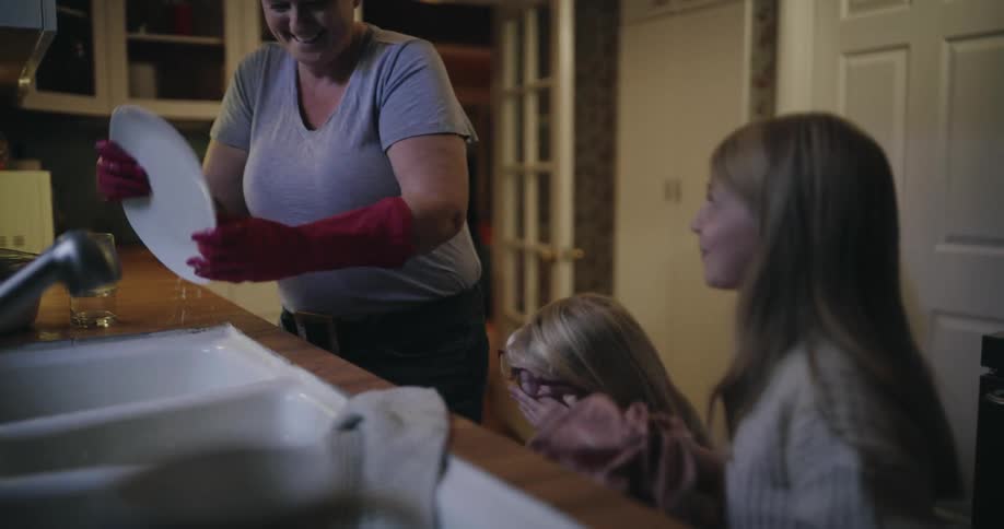 An adult woman wearing red rubber gloves washes a white plate in a kitchen sink while two young girls watch the woman hands the wet plate to the younger girl who playfully tries to dry it with a cloth