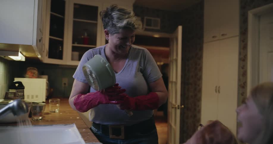 A woman wearing red rubber gloves and a grey shirt playfully scares two young girls in a kitchen while holding a pot and a wooden spoon during a moment of family fun