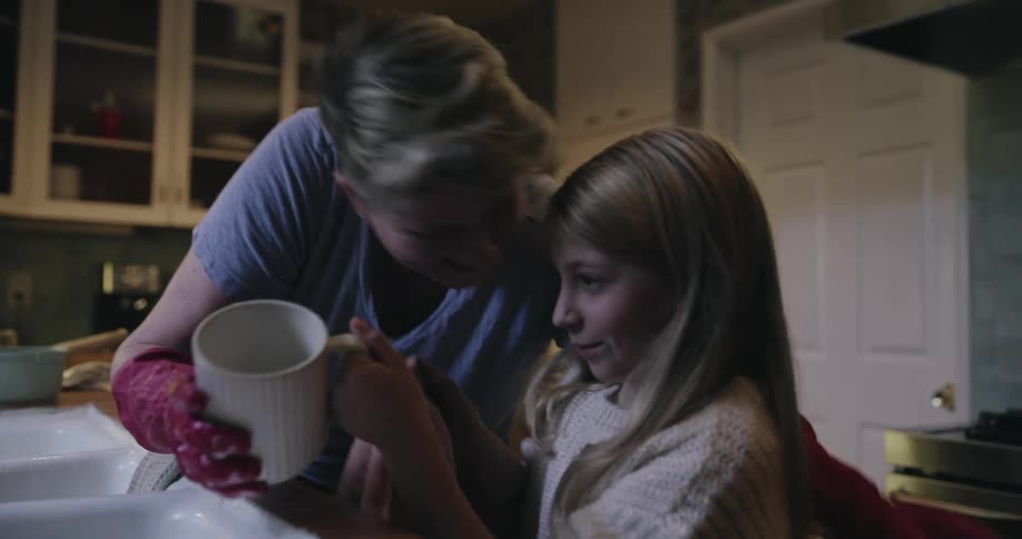 In a kitchen a woman wearing red gloves washes a mug and gives a young girl a kiss on the forehead and the mug the girl begins to dry it with a towel while they both smile during the shared chore