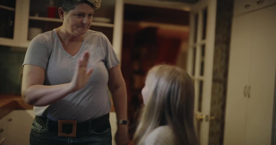 A woman in a grey shirt and blue jeans finishes cleaning a kitchen counter with a cloth before giving a high five to a young girl and walking out of the room together through a doorway