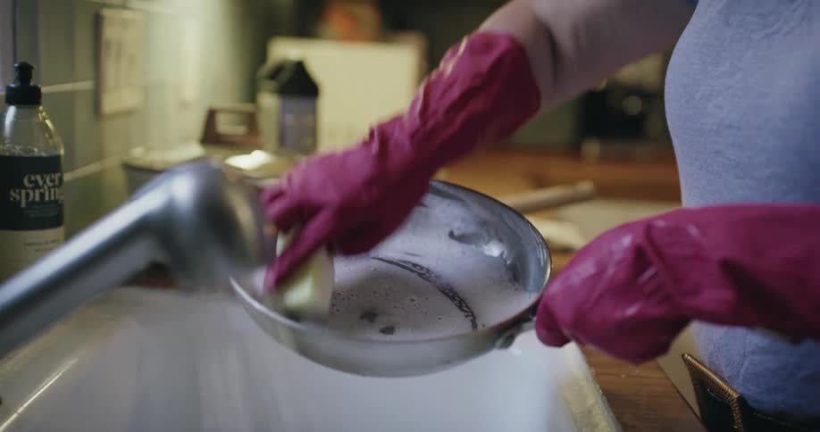 A woman with short grey hair wearing pink rubber gloves scrubs a metal frying pan with a soapy sponge in a kitchen sink