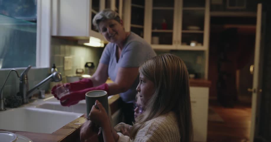 A woman wearing pink rubber gloves washes dishes in a kitchen sink while a young girl stands beside her drying a dark mug with a cloth towel