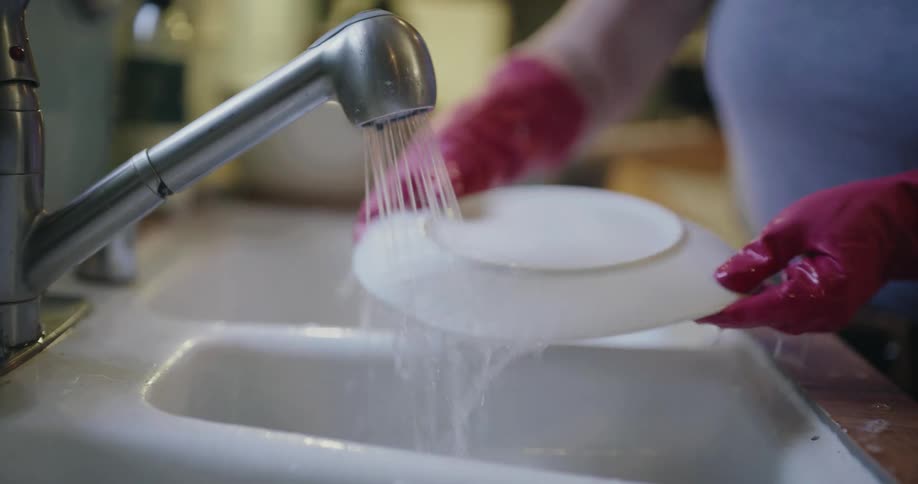 A young woman rinsing a white dish and handing it to her son as he wipes it with a napkin with another young girl standing beside them in the kitchen at midday