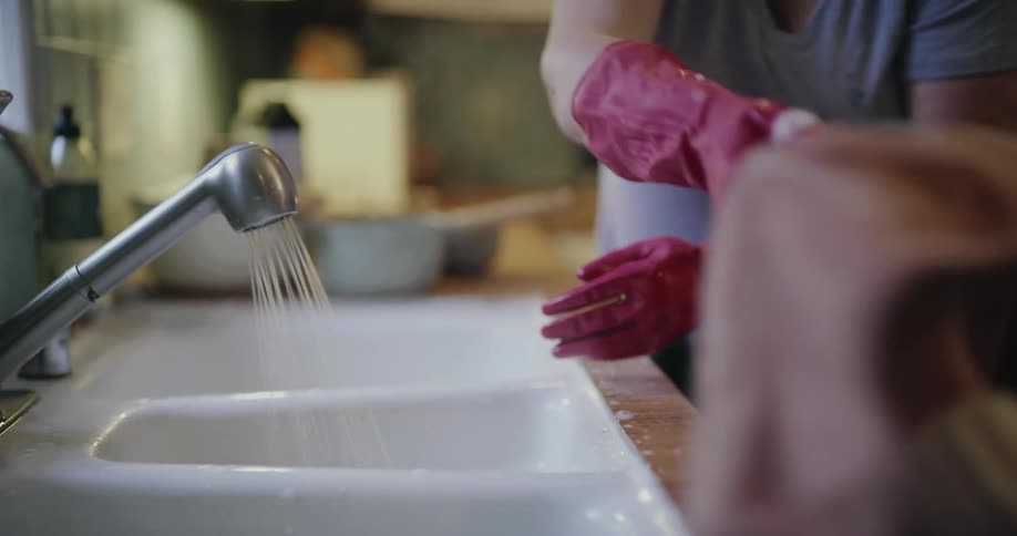 A smiling woman wearing red rubber gloves washes a grey mug in a white kitchen sink before rinsing a white textured mug and handing it to a young blonde girl standing beside the wooden counter