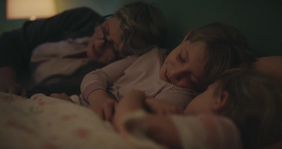 Adult sleeps beside two children in a bed under a blanket indoors