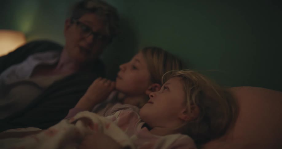 A woman with glasses reads a book to two young girls in a dimly lit bedroom at night before they settle down to sleep under a patterned blanket