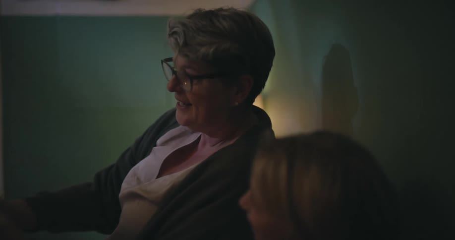 A woman wearing glasses and a grey cardigan sits on a bed and reads a book aloud to two young girls tucked under a floral blanket in a dimly lit bedroom at night