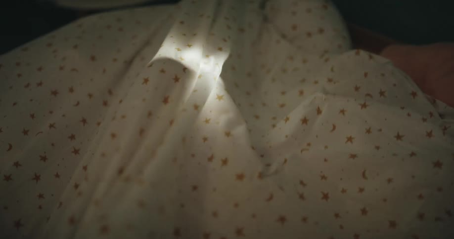 Two children wrapped together under a blanket in a dark room at night illuminated by the soft glow of a flashlight beneath the blanket