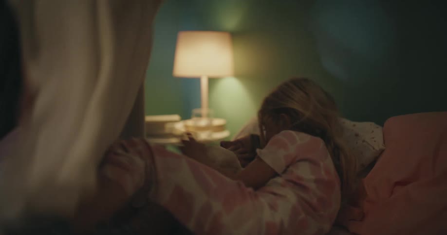 A close view of two young sisters sleeping together in a bed at night while a parent gently tucks them in under a blanket in a dimly lit bedroom
