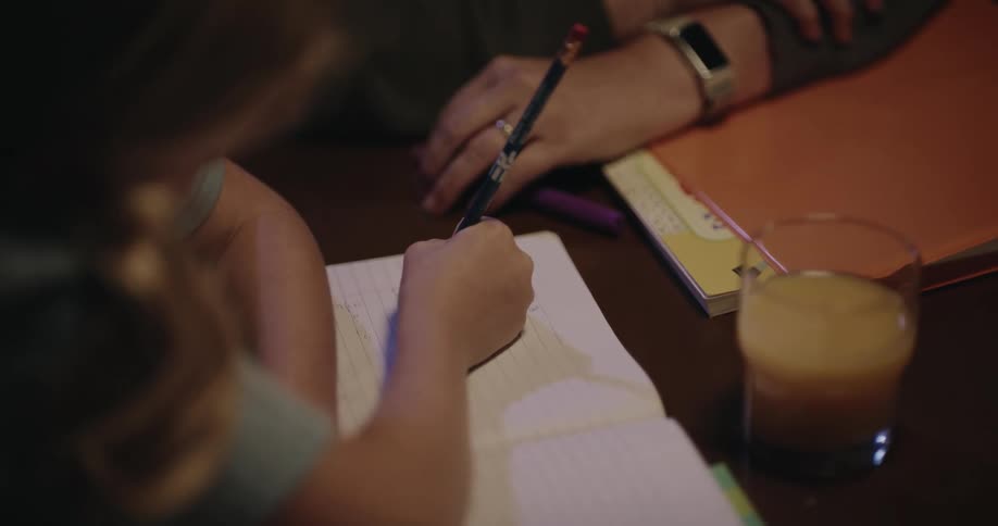 Female child sitting at a table writing on a notebook with an adult woman next to her talking indoors during night time