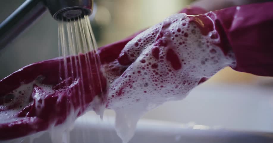 A person wearing pink rubber gloves generates a thick lather using a sponge at a kitchen sink they then rinse the soap suds off the gloves under a steady stream of running water