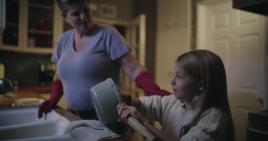 In a softly lit kitchen a woman wearing red gloves washes dishes while a young girl stands beside her drying a pot