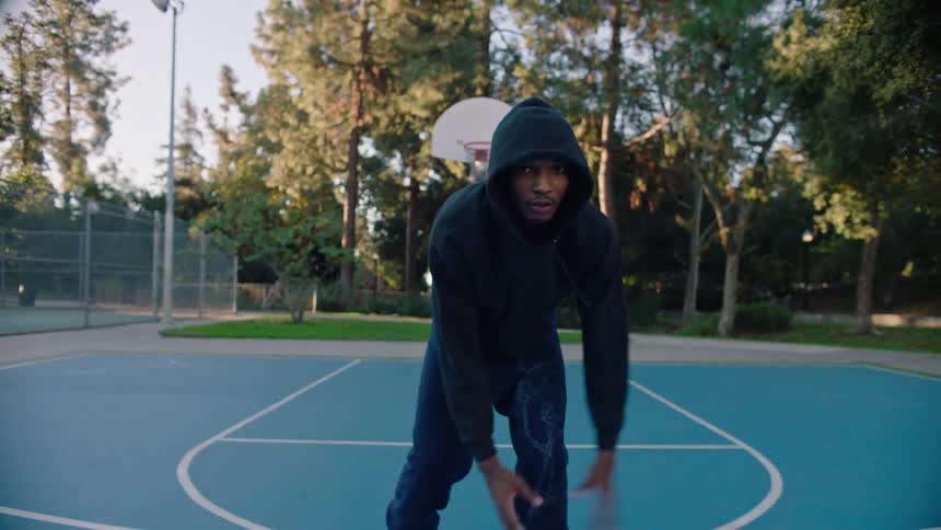A man in a black hooded sweatshirt and dark trousers shoots a basketball on an outdoor blue court surrounded by tall palm trees and lush greenery under a clear sky