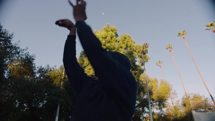 Young adult black man wearing a black hoodie holding a basketball over his head and then throwing it outdoors during dusk or dawn