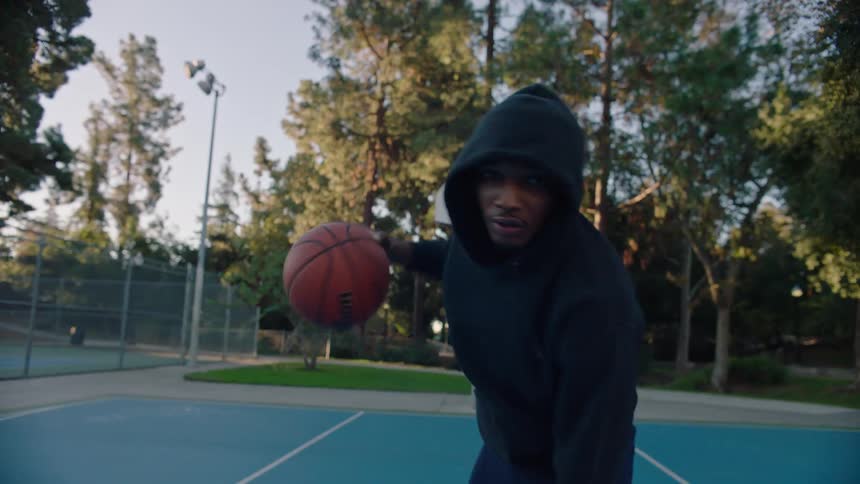 A man wearing a black hoodie looks directly into the camera while holding a basketball on an outdoor court with tall green trees and a clear sky in the background