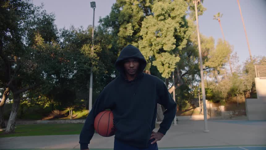 A man wearing a black hoodie stands on an outdoor basketball court holding a basketball under his arm with green trees and a hoop in the background during the late afternoon