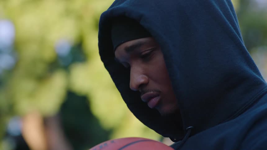 A close view of a man wearing a black hoodie looking down thoughtfully while holding a basketball on an outdoor court with blurred green trees in the background