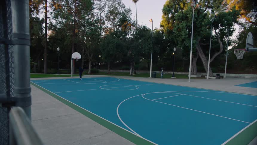 Person plays basketball alone on an outdoor court surrounded by trees