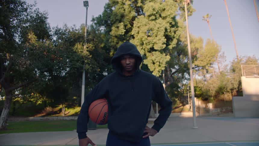 A man wearing a black hooded sweatshirt stands on an outdoor basketball court holding a basketball under his arm with trees and tall palm trees in the background