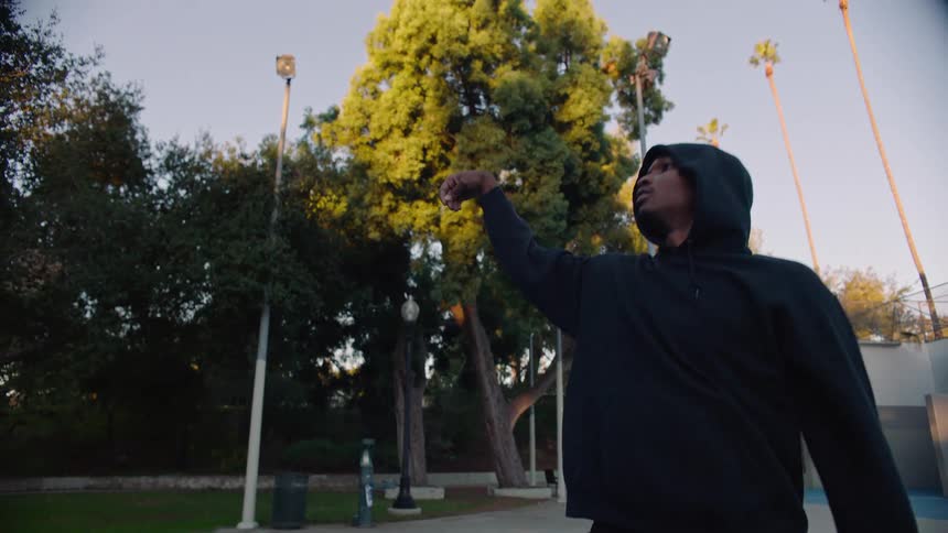 A man wearing a black hoodie shoots a basketball on an outdoor court surrounded by tall palm trees and lush greenery during the late afternoon