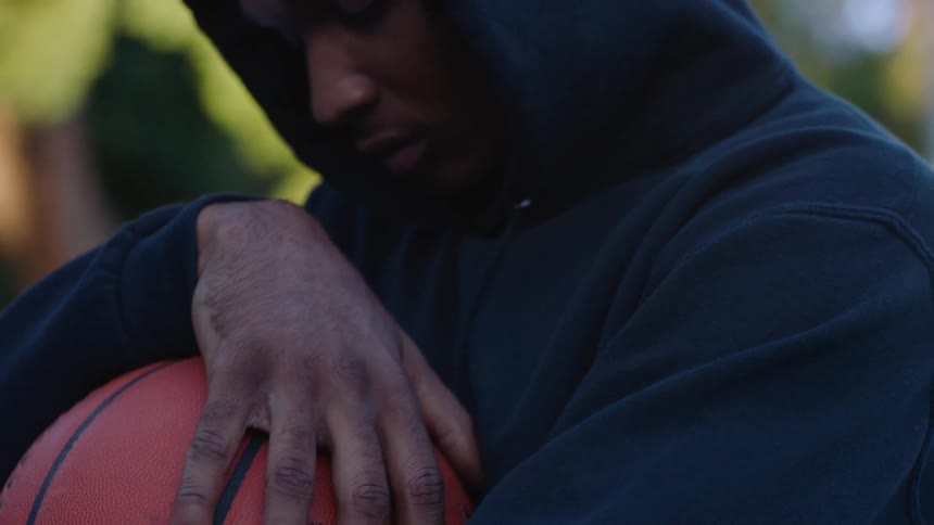 A young man wearing a dark hoodie stands outside cradling a basketball close to his chest he leans his head down toward the ball with a somber or pensive expression
