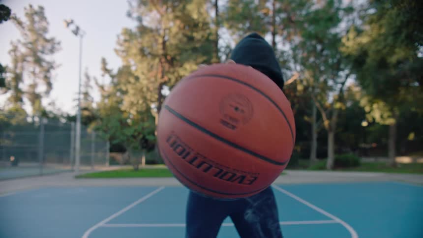 Young black man wearing a hoodie holding and moving around a basketball on a basketball court at a park outdoors during dusk or dawn