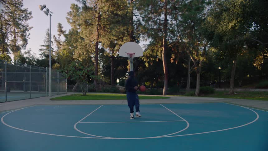 A person wearing a dark hoodie and pants dribbles a basketball on a blue outdoor court before jumping to take a shot at the hoop surrounded by tall trees and a fence under clear daylight