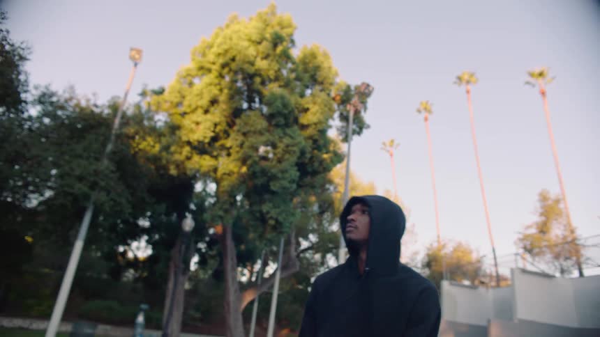 A young man wearing a black hoodie stands on an outdoor basketball court and tosses a ball into the air against a backdrop of tall palm trees and green foliage at dusk