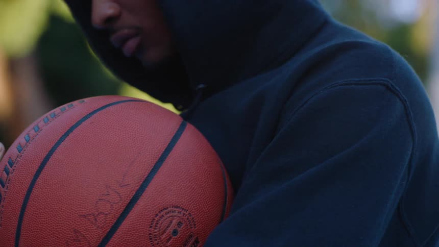 A young man wearing a black hoodie standing on a basketball court at midday holding a basketball in his hands under the bright sun