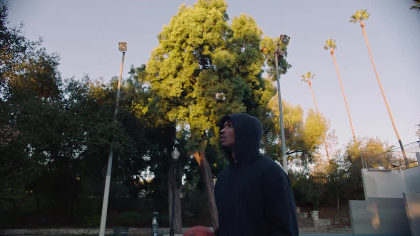 Young adult black man wearing a hoodie throwing a basketball at a park outdoors during dusk or dawn