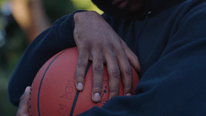 A man wearing a dark hooded sweatshirt looks down thoughtfully while holding an orange basketball against his chest with both hands in a sunlit outdoor environment