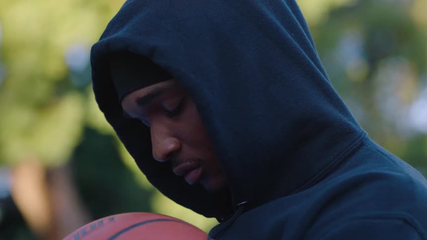 Young black male wearing a bandanna and a hoodie holding a basketball outdoors with a contemplative facial expression