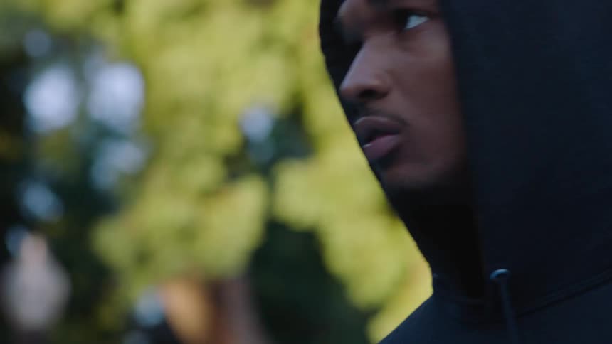 Young adult black man wearing a black hoodie and bandanna dancing outdoors with trees on the background during day time