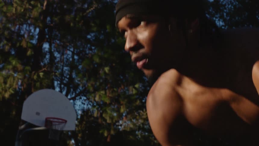 A shirtless young man with a black headwrap focuses intensely while playing basketball on an outdoor court breathing heavily during a closeup profile shot before the camera cuts to a quick movement of him dribbling the ball on the blue ground