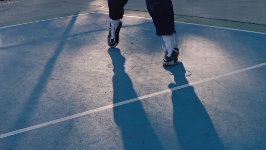A low angle view of a person dribbling a basketball on a blue outdoor court with long shadows cast on the ground during the late afternoon