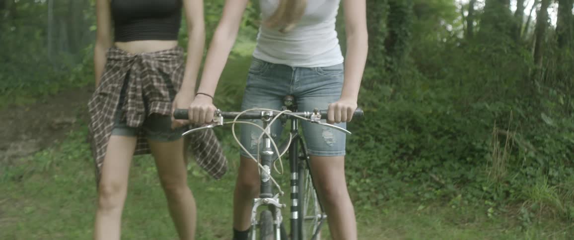 One woman rides a bike and one walks next to her through the woods on a sunny day