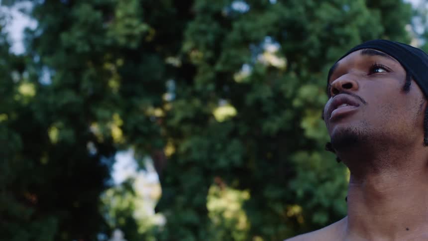 A shirtless young man with a black headwrap jumps and shoots a basketball outdoors against a backdrop of green trees during a sunny day in a closeup lowangle shot